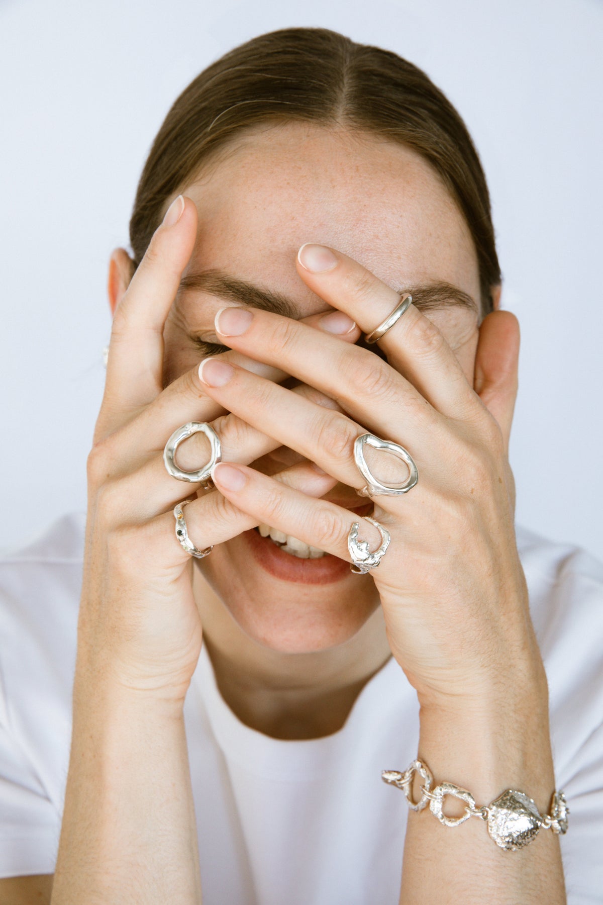 A close-up lifestyle shot of a woman covering her eyes with her hands, wearing multiple silver rings and a silver bracelet against a clean white background.